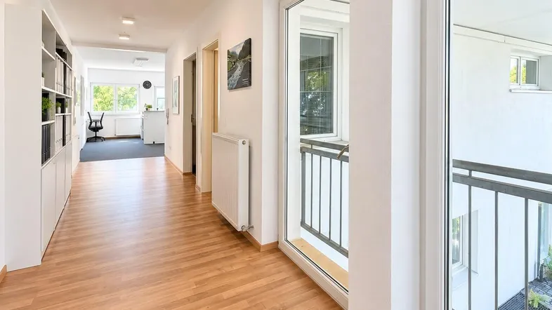 Heller Flur mit Holzfußboden, weißen Wänden und einem Blick auf den Außenbereich sowie ein Büro im Hintergrund.