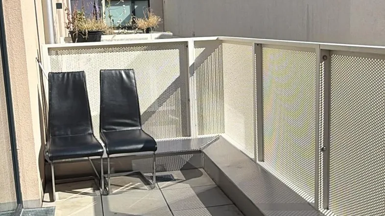Schmaler Balkon mit Fliesenboden und zwei Stühlen, umgeben von modernen Gebäuden.