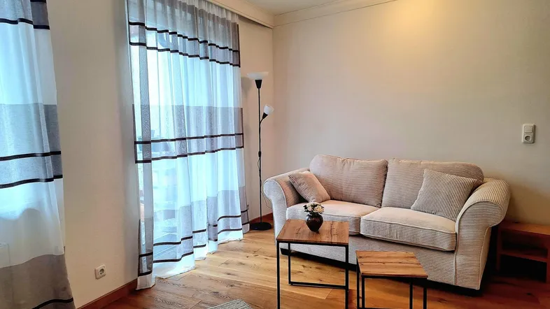 Helles Wohnzimmer mit beigem Sofa, Holzboden und Zugang zu einem Balkon oder einer Terrasse.