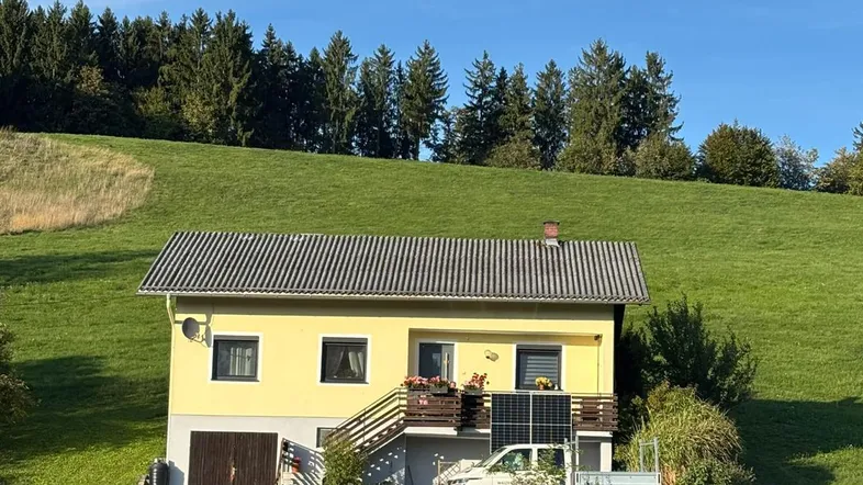 Gelbes Einfamilienhaus auf einem grünen Hügel mit Wald im Hintergrund unter blauem Himmel.
