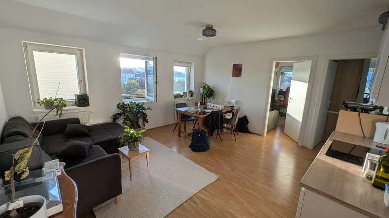 Helles Wohn- und Esszimmer mit Parkettboden, bequemer Couch und Esstisch, sowie einem kleinen Küchenbereich.