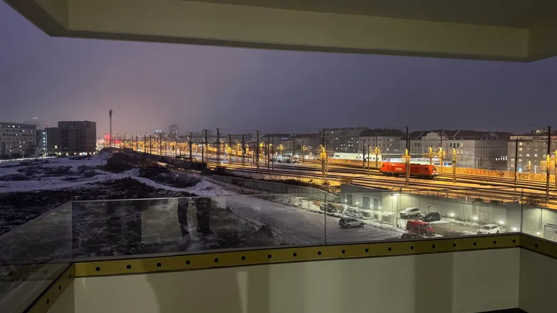 Blick von einem modernen Balkon mit Glasgeländer auf die beleuchteten Bahngleise und die verschneite Stadt bei Nacht.