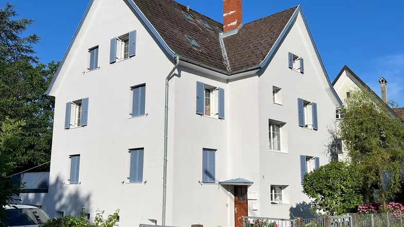 Weißes Wohnhaus mit blauen Fensterläden und Satteldach unter klarem Himmel.