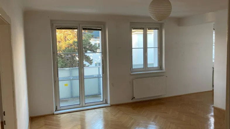Helles Wohnzimmer mit Fischgrätparkettboden, großen Fenstern und Zugang zu einem Balkon.