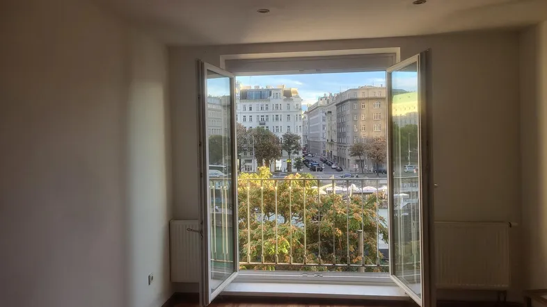 Helles Zimmer mit Holzboden und Blick auf die Stadt durch die offene Fenstertür.