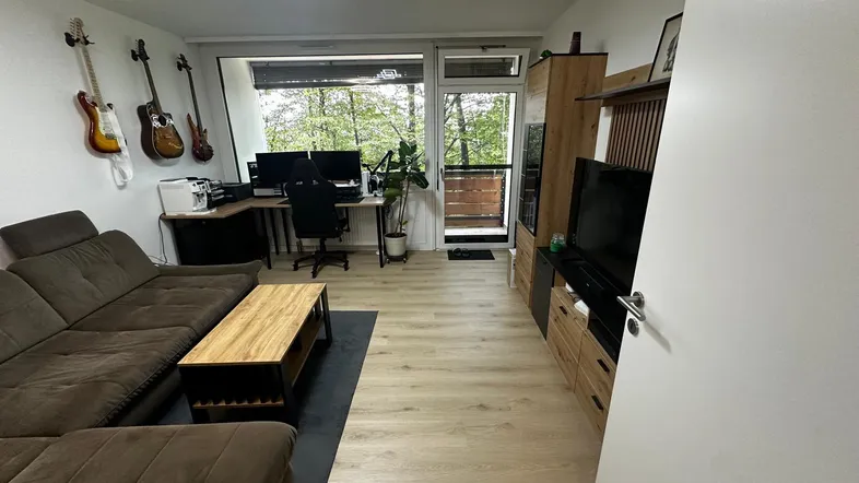 Helles Wohnzimmer mit großem Sofa, Arbeitsbereich und Zugang zum Balkon mit Blick ins Grüne.