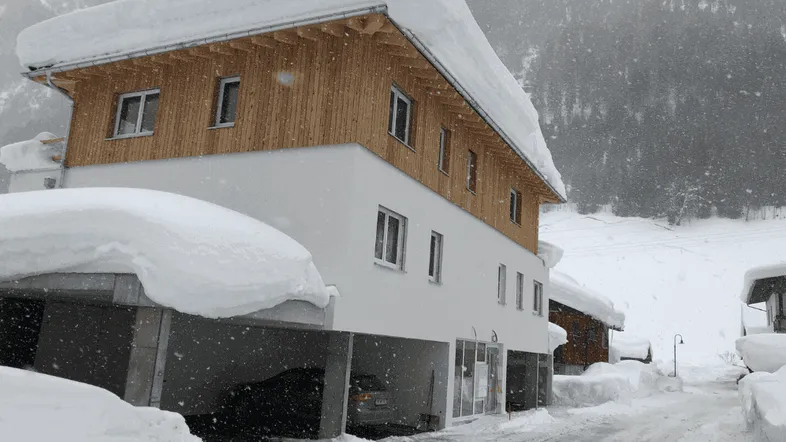 Modernes Gebäude mit Holzfassade und integriertem Carport, umgeben von tiefem Schnee in den Bergen.