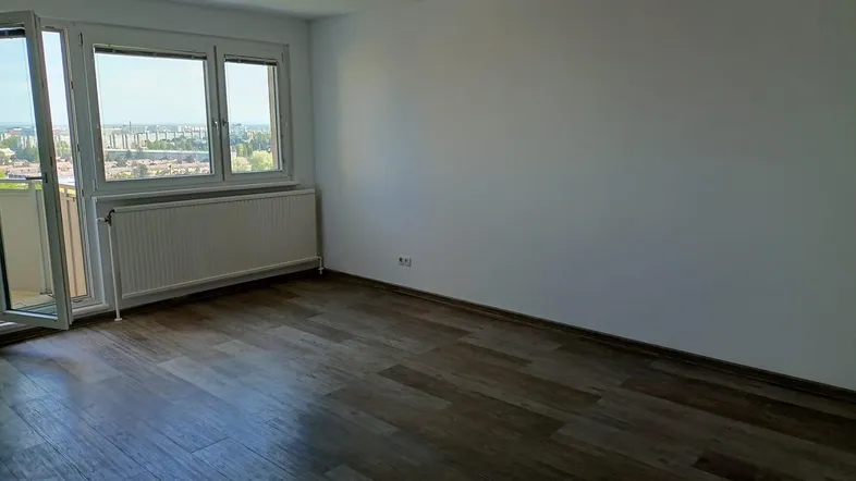 Helles Zimmer mit Laminatboden, Heizkörper und Zugang zu einem Balkon mit Stadtblick.