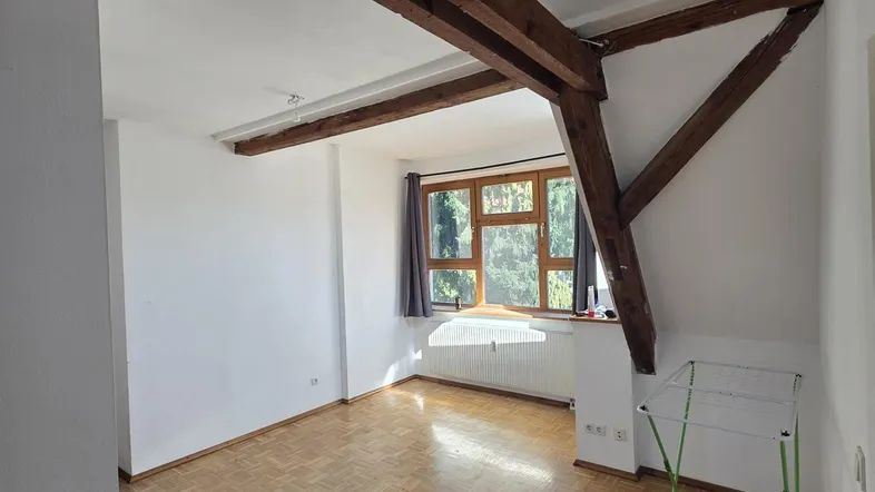 Helles Zimmer im Dachgeschoss mit sichtbaren Holzbalken, Parkettboden und großem Fenster.