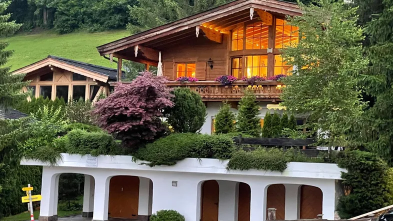 Traditionelles Holzhaus im alpinen Stil mit beleuchteten Fenstern und überdachten Parkplätzen.