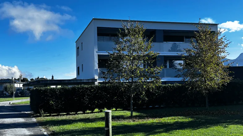 Modernes Wohngebäude mit Balkonen, umgeben von einer grünen Hecke und Rasenfläche unter blauem Himmel.