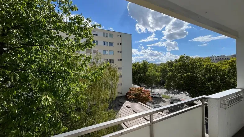 Heller Balkon mit Blick auf Bäume, Straße und umliegende Gebäude unter blauem Himmel.