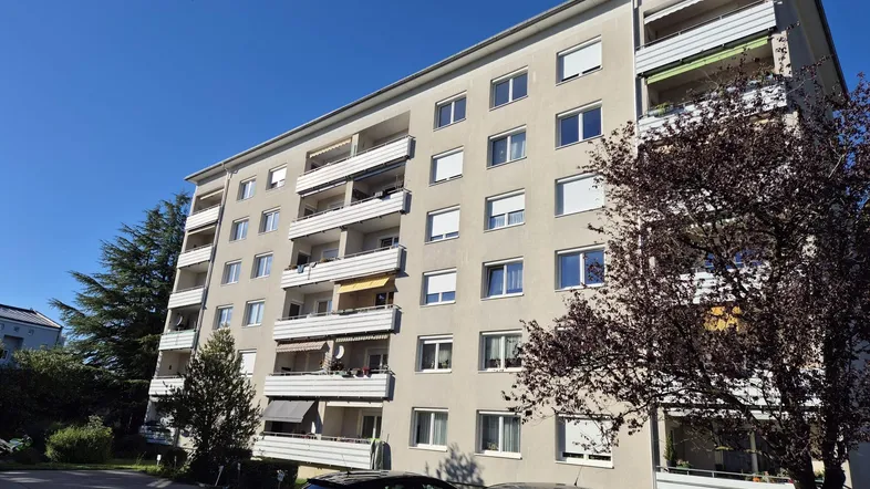 Mehrstöckiges Wohngebäude mit Balkonen und Bäumen im Vordergrund unter blauem Himmel.