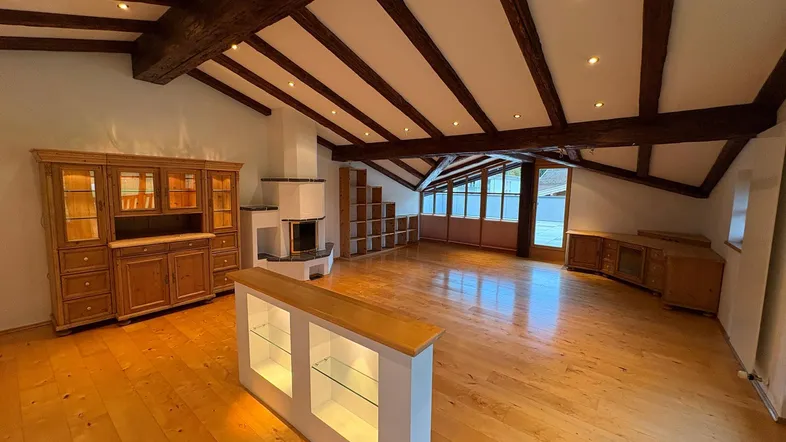 Geräumiges Wohnzimmer mit sichtbaren Holzbalken, Kamin und hellem Holzfußboden unter schräger Decke.