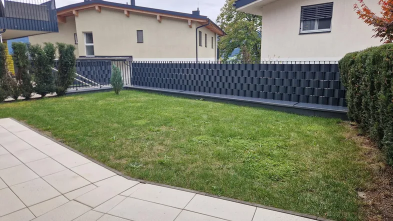 Gepflegter Gartenbereich mit Rasenfläche und gepflasterter Terrasse, umgeben von einer modernen Zaunanlage.
