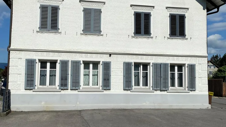 Traditionelles Gebäude mit weißer Fassade und grauen Fensterläden unter blauem Himmel.