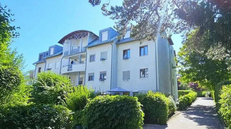 Mehrfamilienhaus mit Mansarddach, Balkonen und gepflegter Grünanlage unter blauem Himmel.