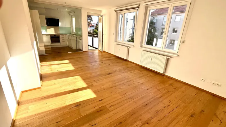 Helles Wohn-Esszimmer mit Holzboden und Zugang zu einer modernen Küche und einem Balkon.