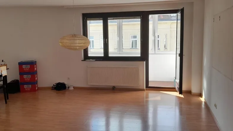 Helles Wohnzimmer mit Parkettboden, großem Fenster und Zugang zu einem kleinen Balkon.