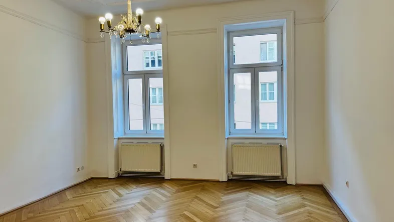 Helles Zimmer mit Fischgrätparkett, hohen Fenstern und Stuckdecke, ideal als Wohnzimmer.