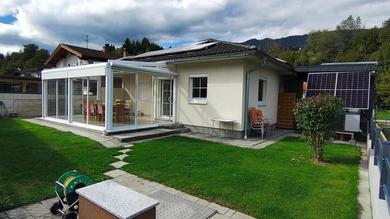 Modernes Haus mit Wintergarten, gepflegtem Rasen und Photovoltaikanlage vor bergiger Kulisse.
