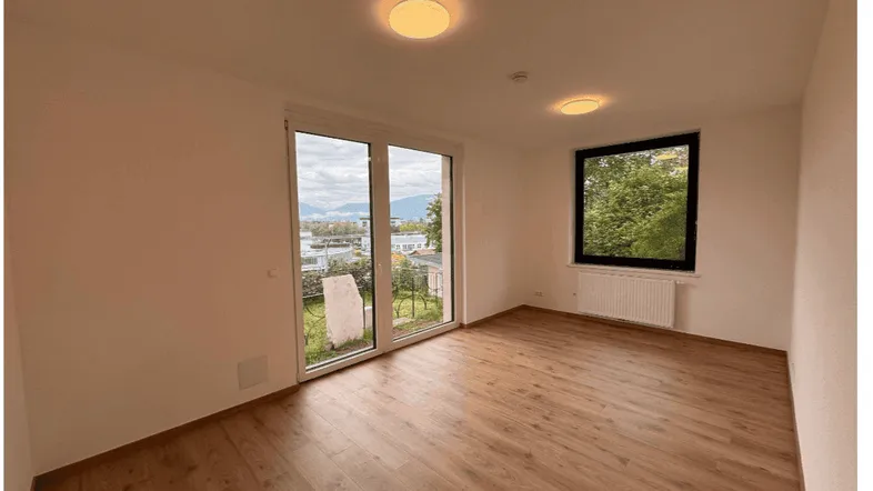 Helles Zimmer mit Holzboden, großen Fenstern und Zugang zu einem kleinen Gartenbereich.