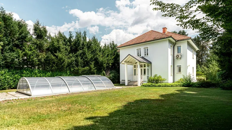 Weißes Einfamilienhaus mit rotem Ziegeldach, großem Garten und überdachtem Swimmingpool unter blauem Himmel.