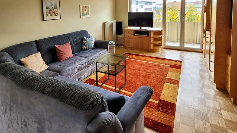 Gemütliches Wohnzimmer mit großem Ecksofa, Teppich und Zugang zum Balkon.