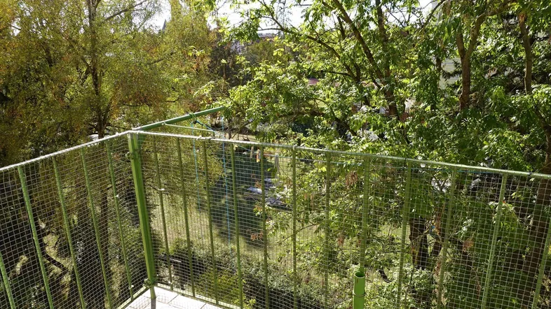 Balkon mit grüner Gitterumzäunung und Blick ins Grüne, umgeben von Bäumen.