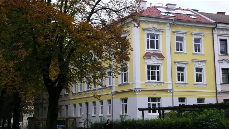 Historisches Gebäude mit gelber Fassade, weißen Fenstern und rotem Dach, umgeben von Bäumen.