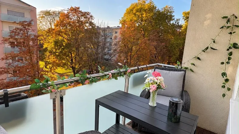 Möblierter Balkon mit Holzboden und Blick auf herbstliche Bäume und umliegende Gebäude.