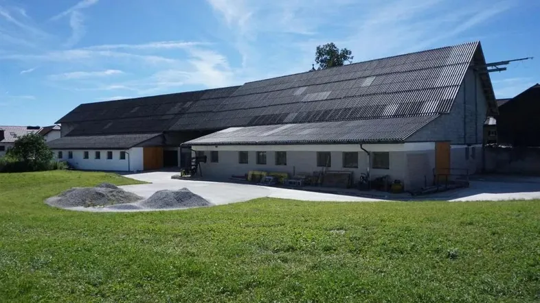 Langes landwirtschaftliches Gebäude mit dunklem Wellblechdach und weiß-grauer Fassade, umgeben von Grünflächen.