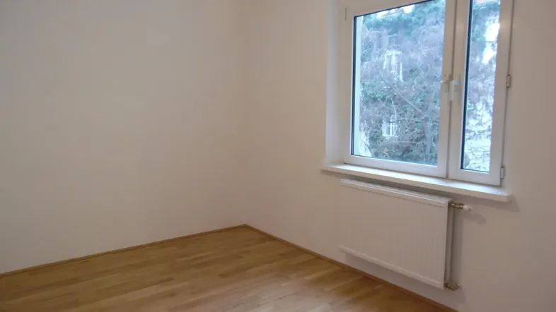 Helles Zimmer mit Parkettboden, großem Fenster und Heizkörper, bietet einen Blick ins Grüne.