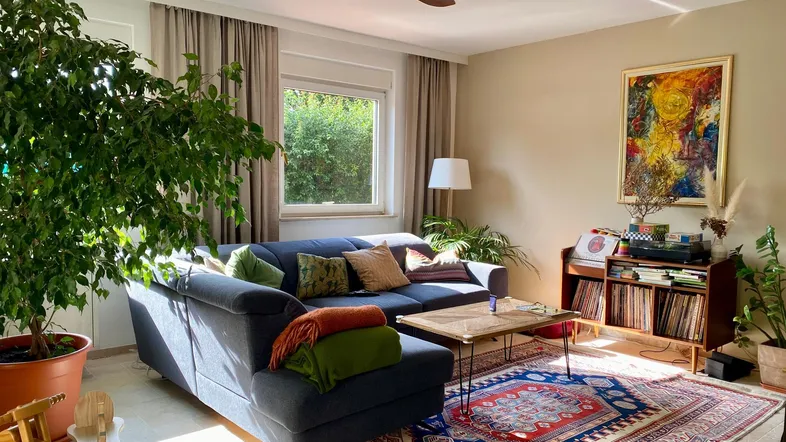 Gemütliches Wohnzimmer mit beigen Wänden, bequemer Couch, Deckenventilator und vielen Pflanzen.