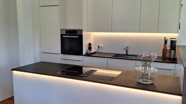 Moderne Küche mit weißen Schränken, dunkler Arbeitsplatte und einer beleuchteten Kücheninsel.