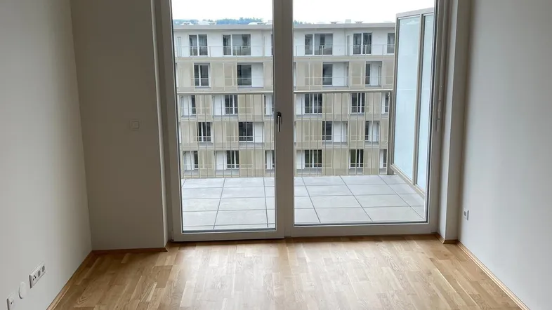 Helles, leeres Zimmer mit Parkettboden und Zugang zu einem großen Balkon mit Blick auf ein gegenüberliegendes Gebäude.