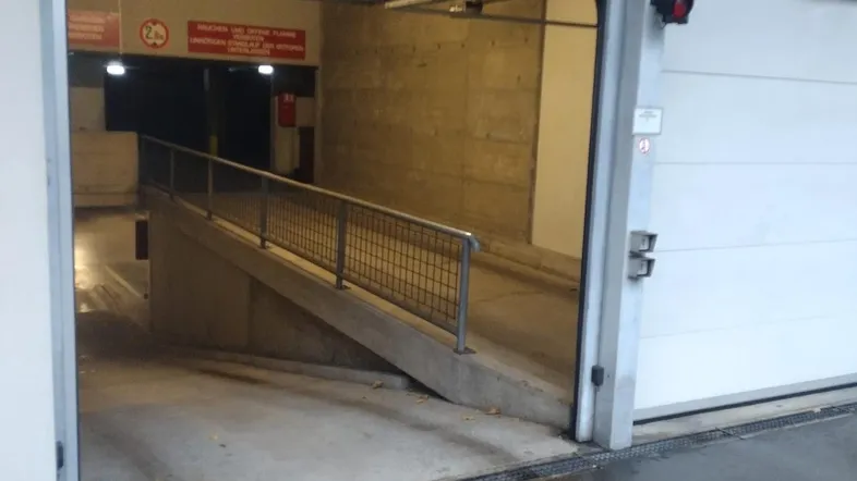 Einfahrt zur Tiefgarage mit Rampe und separatem Fußgängerzugang, beleuchtet und gut sichtbar.