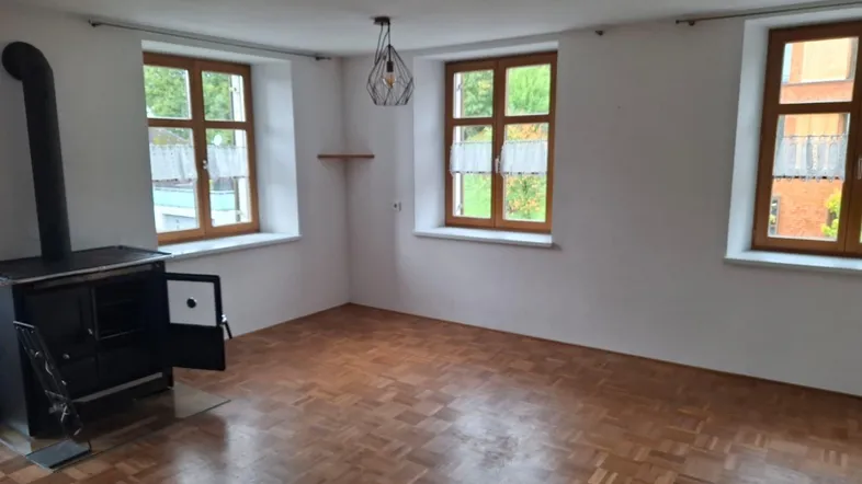 Heller Wohnraum mit Parkettboden, Holzfenstern und einem traditionellen Holzofen als Heizquelle.