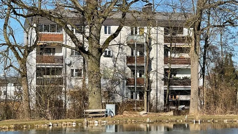 Mehrfamilienhaus am Ufer eines zugefrorenen Teichs, umgeben von hohen Bäumen unter blauem Himmel.