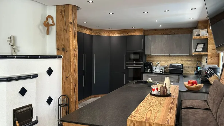 Moderne Küche mit dunklen Fronten, Holzelementen und einem traditionellen Kachelofen als Blickfang.
