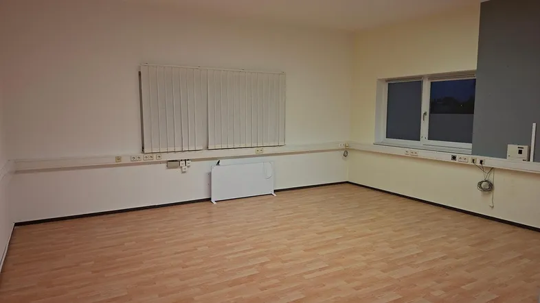 Leerer Büroraum mit Laminatboden, großen Fenstern und Deckenbeleuchtung, bereit zur Nutzung.