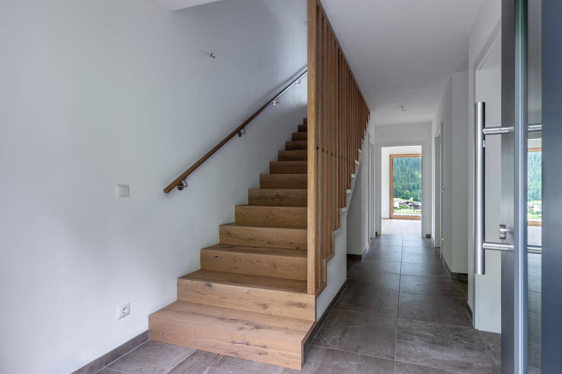 Heller Flur mit Holztreppe, modernen Geländern und Blick ins Grüne durch ein Fenster.