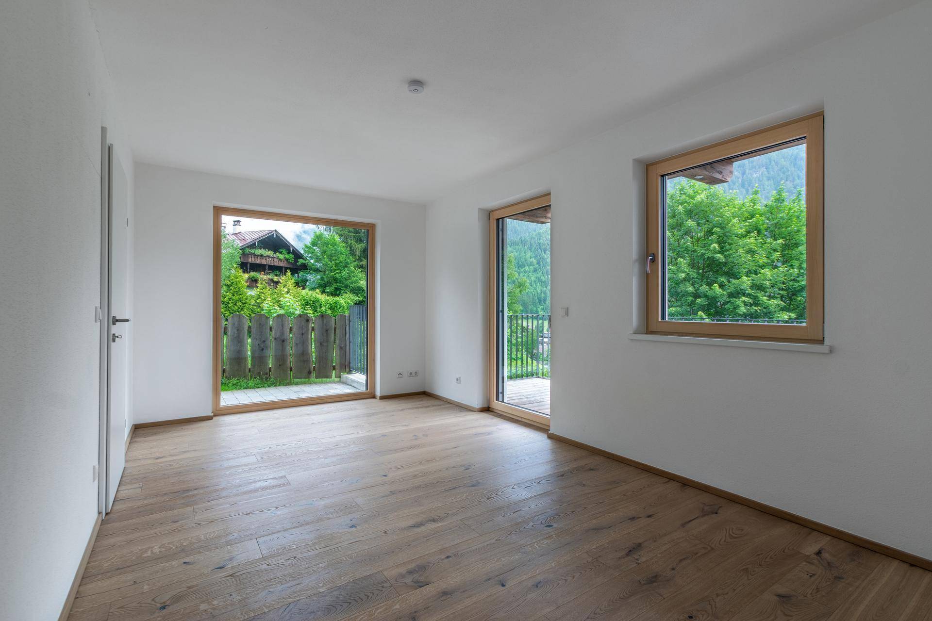 Helles Schlafzimmer mit Holzboden und großen Fenstern, die Zugang zu einem Außenbereich mit viel Grün bieten.