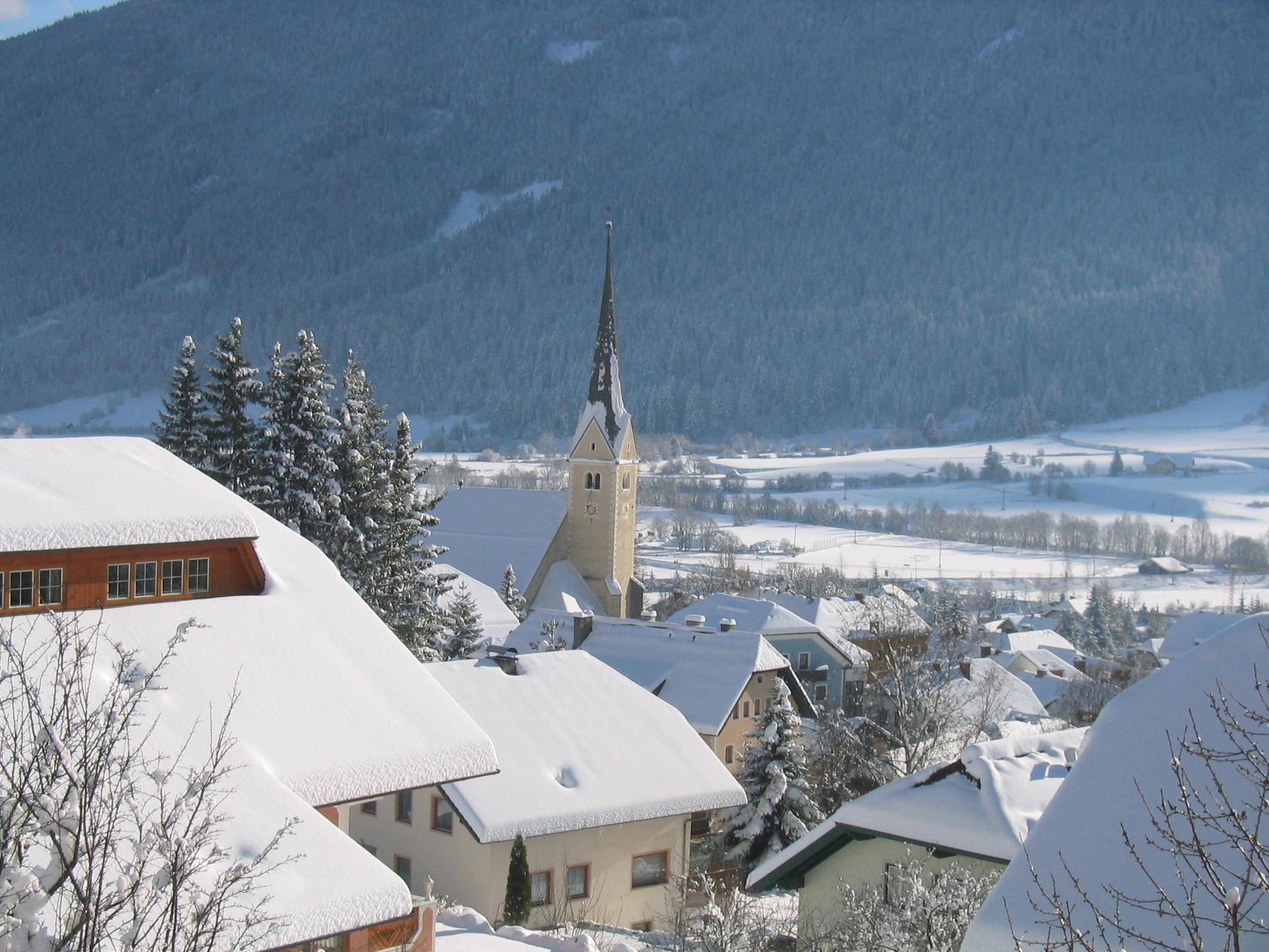 Winterlandschaft mit verschneitem Dorf, Kirche und Bergen im Hintergrund.