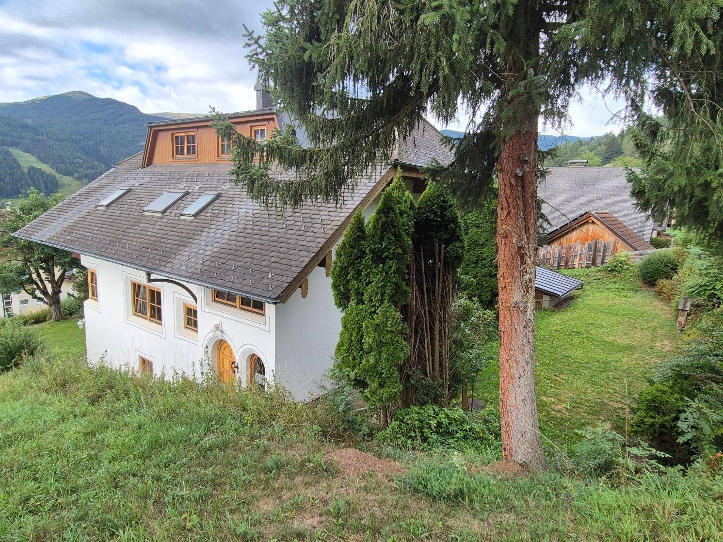 Traditionelles Haus mit Satteldach und Gauben, umgeben von grüner Landschaft und Bäumen.