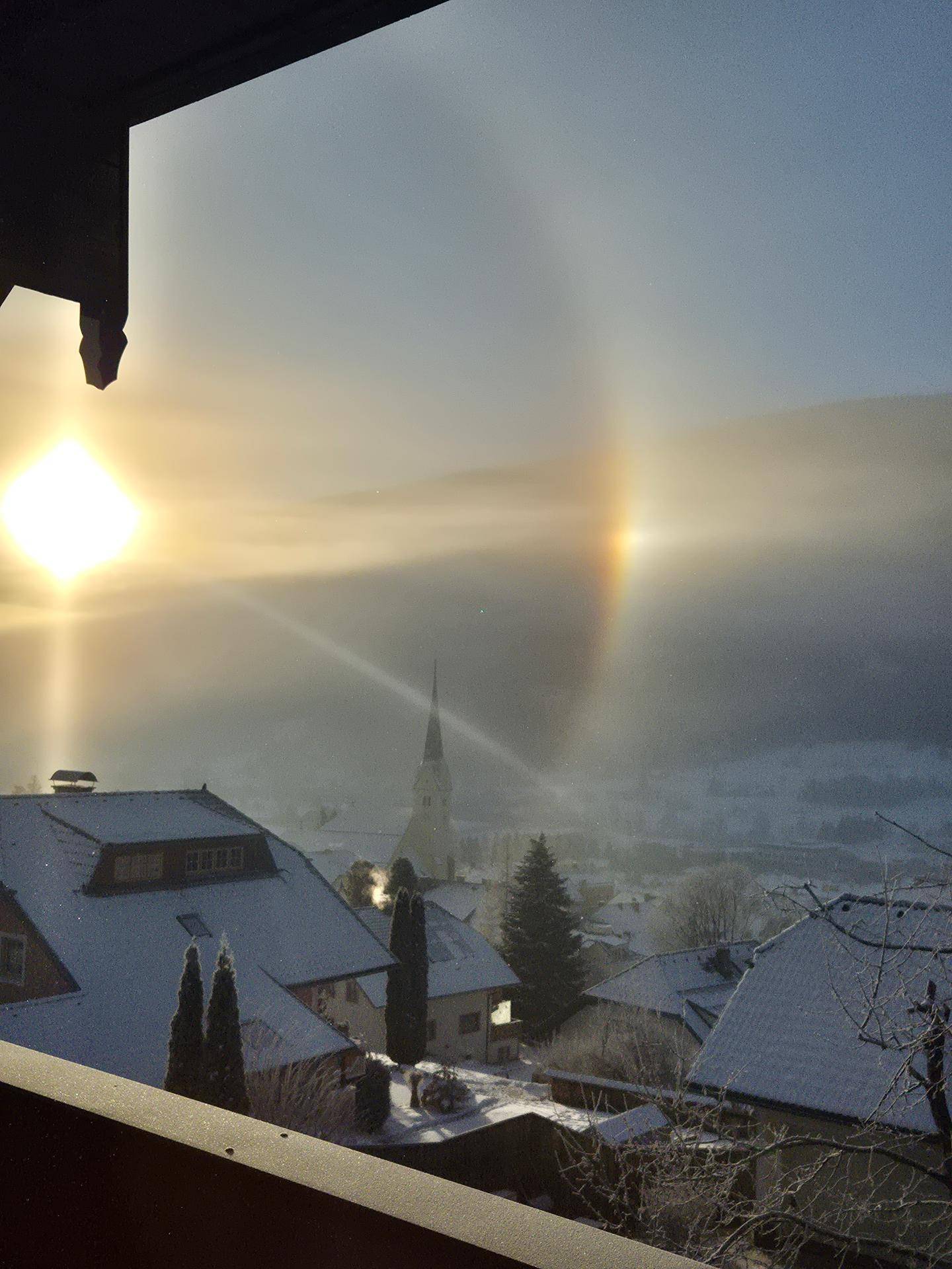 Winterlicher Sonnenaufgang über einem verschneiten Dorf mit Kirche und beeindruckendem Lichtspiel.
