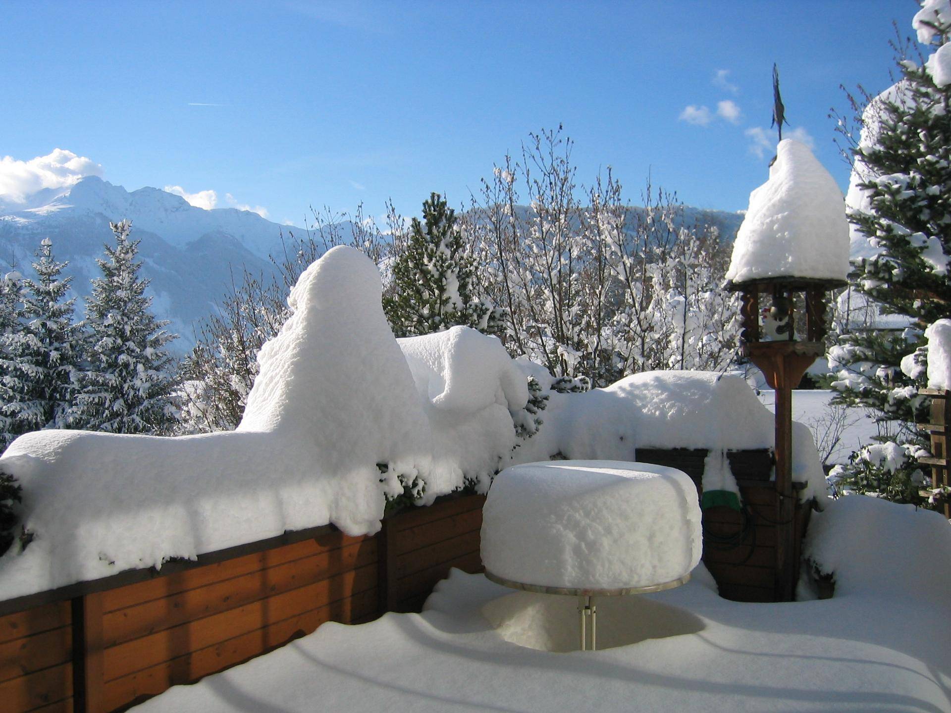 Winterlicher Balkon mit tiefem Schnee, umgeben von verschneiten Bäumen und Bergblick.