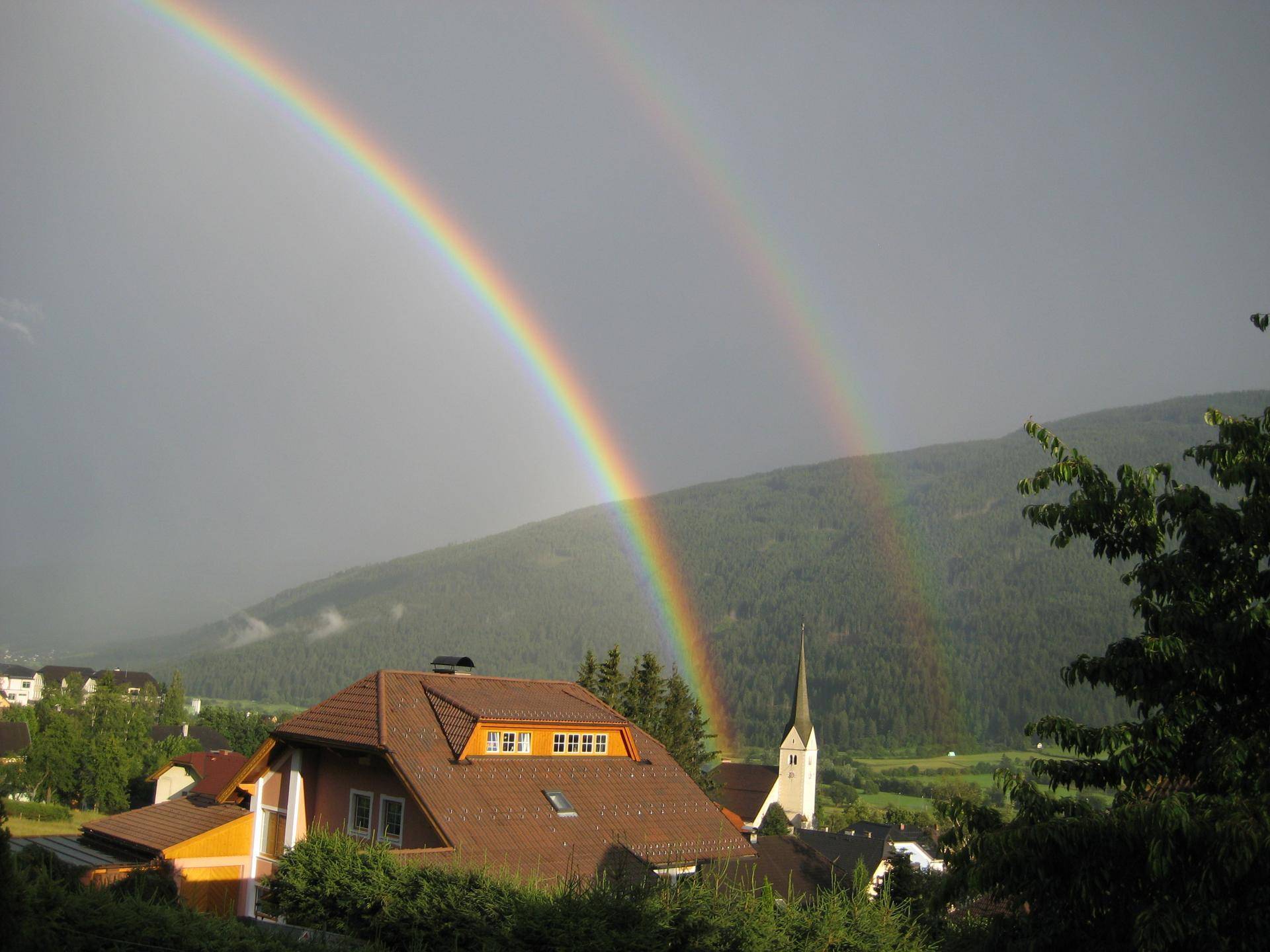 Malerischer Ausblick auf ein Dorf mit Kirche und einem doppelten Regenbogen über den Bergen.