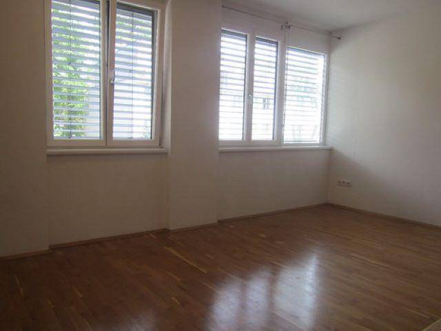 Helles, leeres Zimmer mit Holzparkettboden und großen Fenstern mit Jalousien.