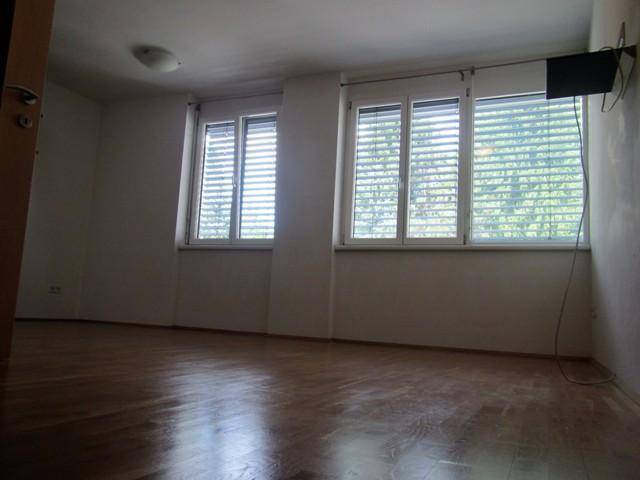 Geräumiges, helles Zimmer mit Holzparkettboden und mehreren Fenstern mit Jalousien.